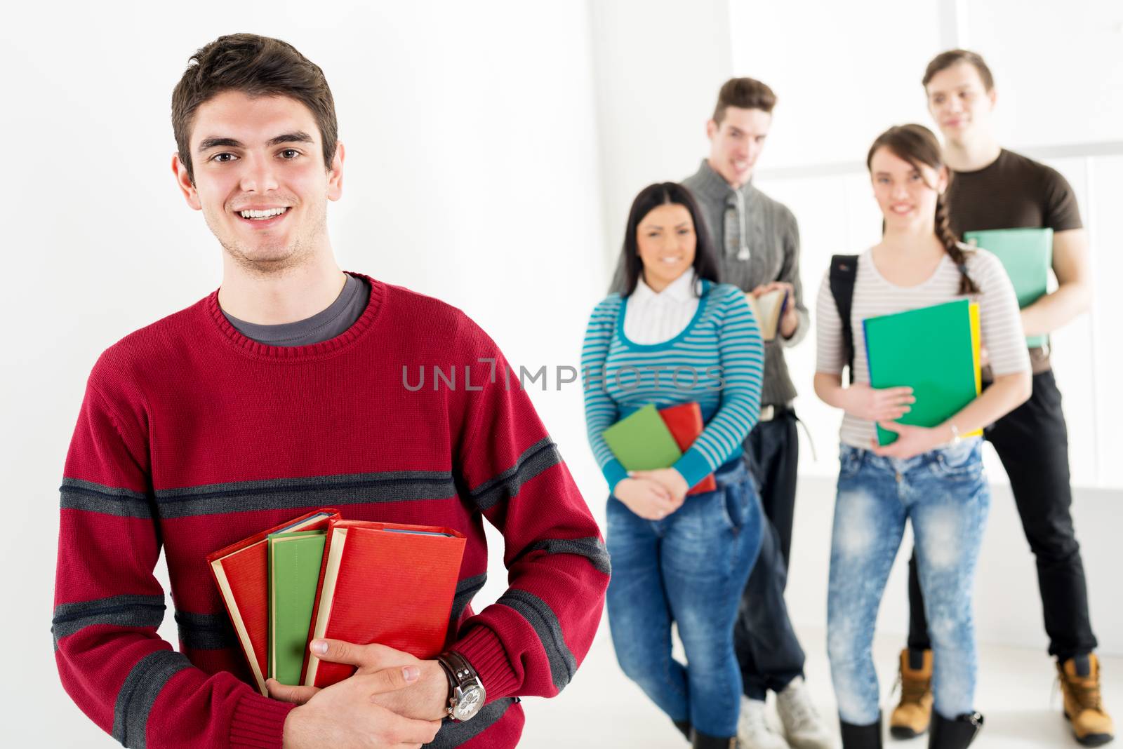 A beautiful smiling young man with books is standing in the foreground. A happy group of his friends is behind him. Looking at camera.