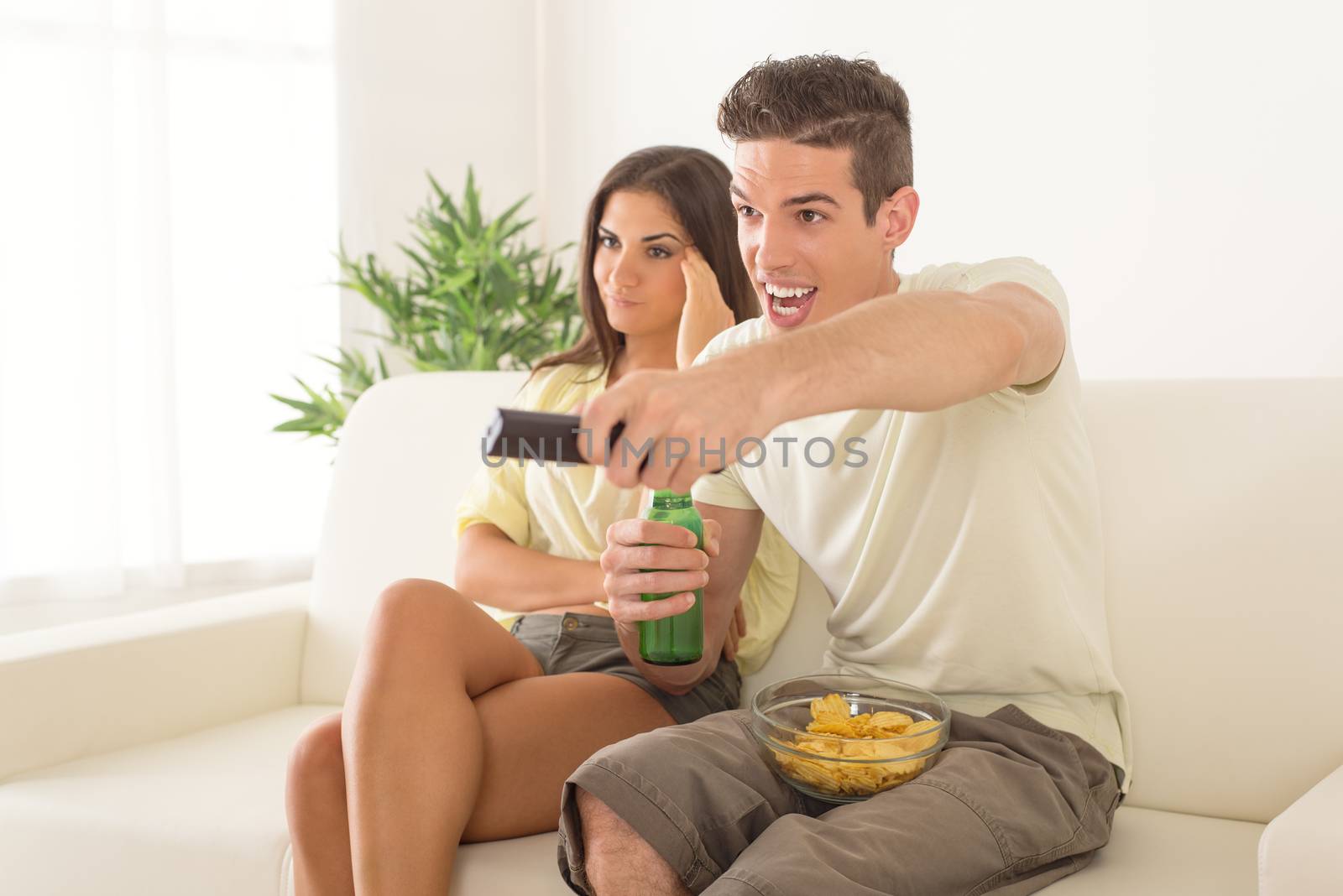 Young man sitting on sofa and watching football on tv. His girlfriend is behind, frowning because and bored.