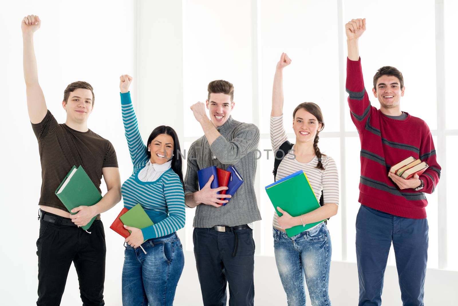 Group of cheerful students with books standing with arms raised in a fist in school hall and looking at camera.