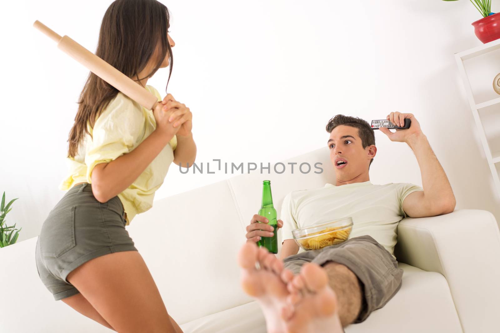 Young man sitting on sofa and watching tv. His wife is angry and standing with rolling pin.