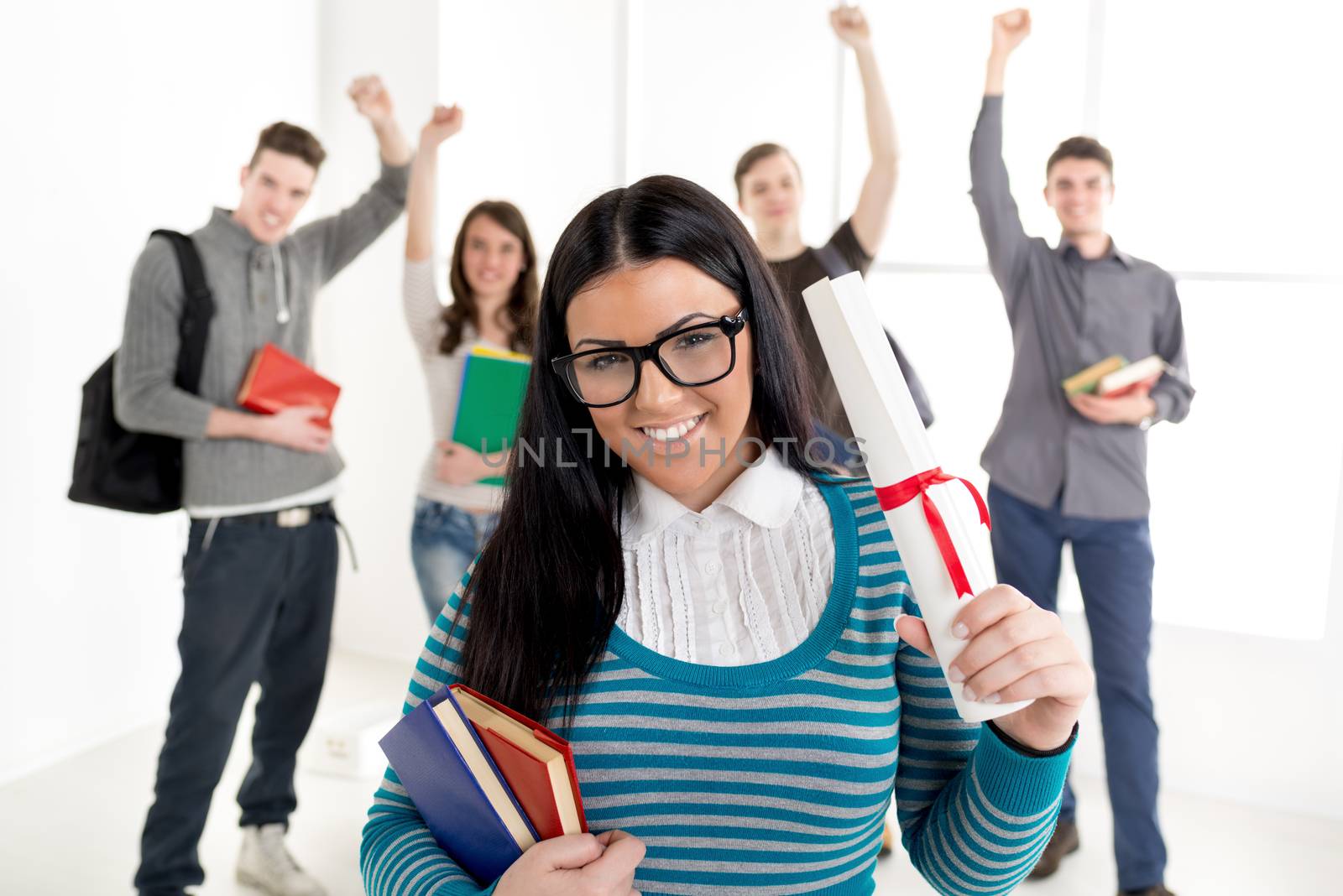 A beautiful smiling young student girl with diploma is standing in the foreground. A happy group of his friends is behind him with arms raised in a fist in the air.
