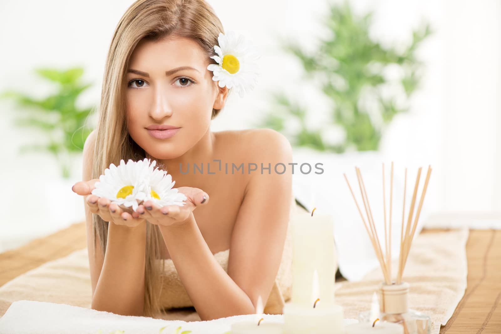 Cute young woman enjoying during a skincare treatment at a spa. She is smiling the camera and holding white flowers in her hands. 