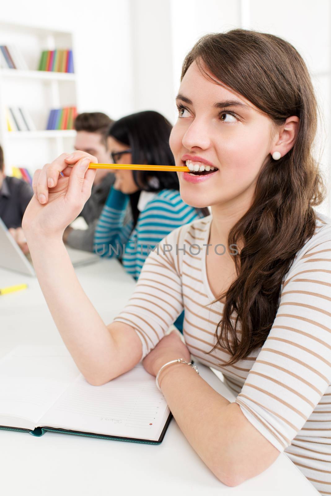 A beautiful smiling girl learning, Biting her pencil and thinking in the foreground. A happy group of her friends is behind him and looking at laptop. 