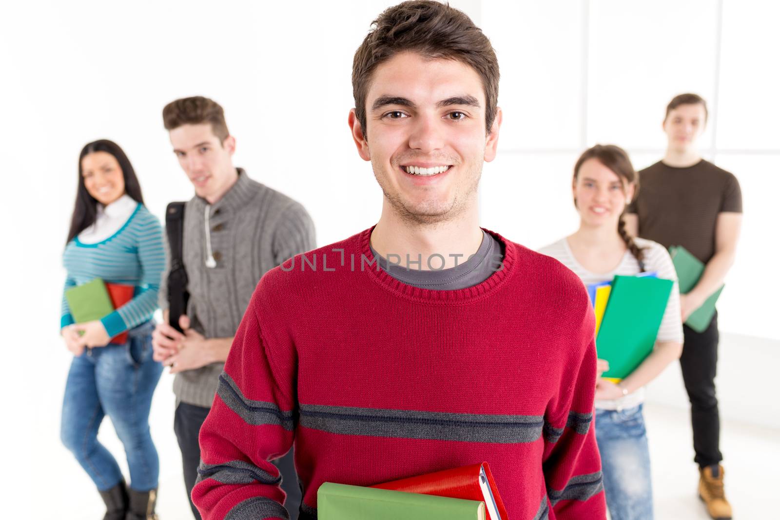 A beautiful smiling young man with books is standing in the foreground. A happy group of his friends is behind him. Looking at camera.