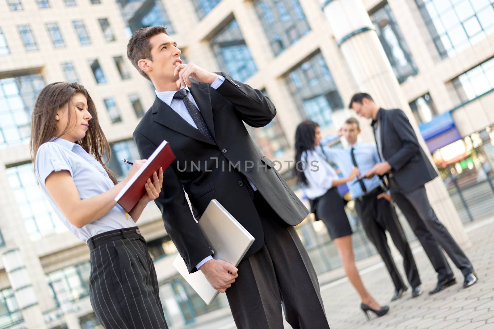 Young businessman and his secretary standing in front of office building.  Businessman carrying in one hand a closed laptop and thinking, and the secretary keeps the planner.