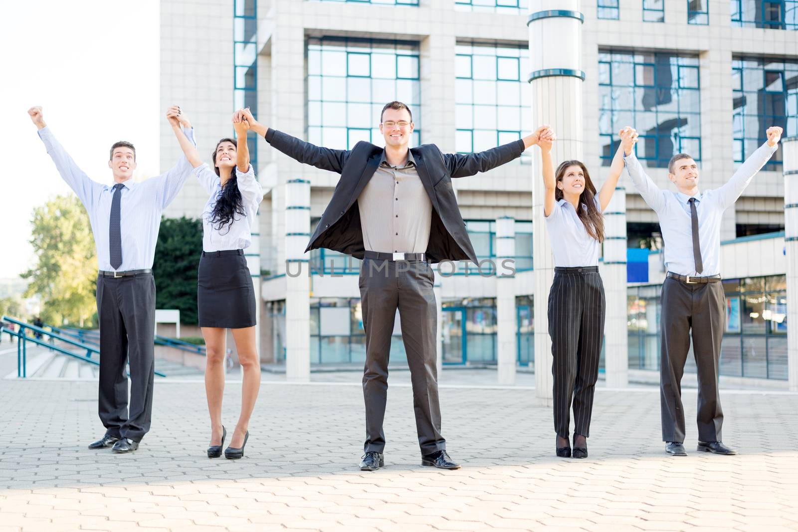 Small group of business people outside their company, holding hands raised celebrate business success.