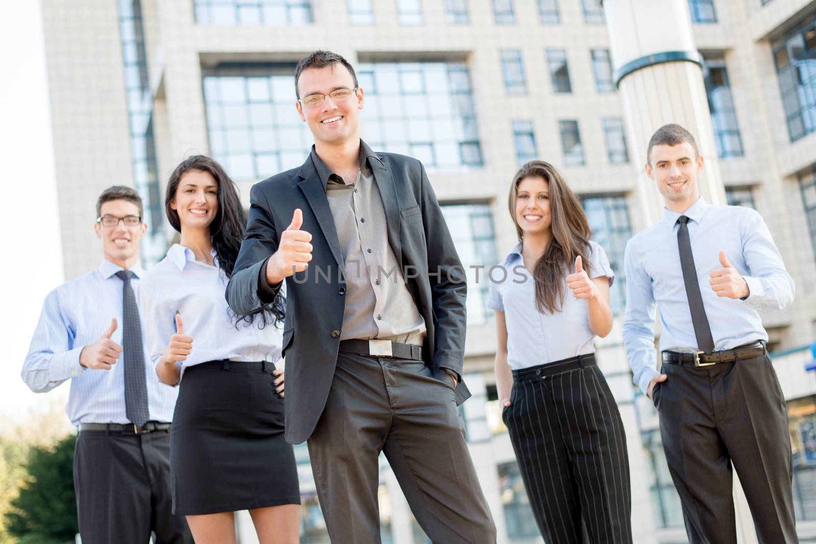 Young business team, elegantly dressed standing outside in front of office building with thumbs up. Looking at camera.