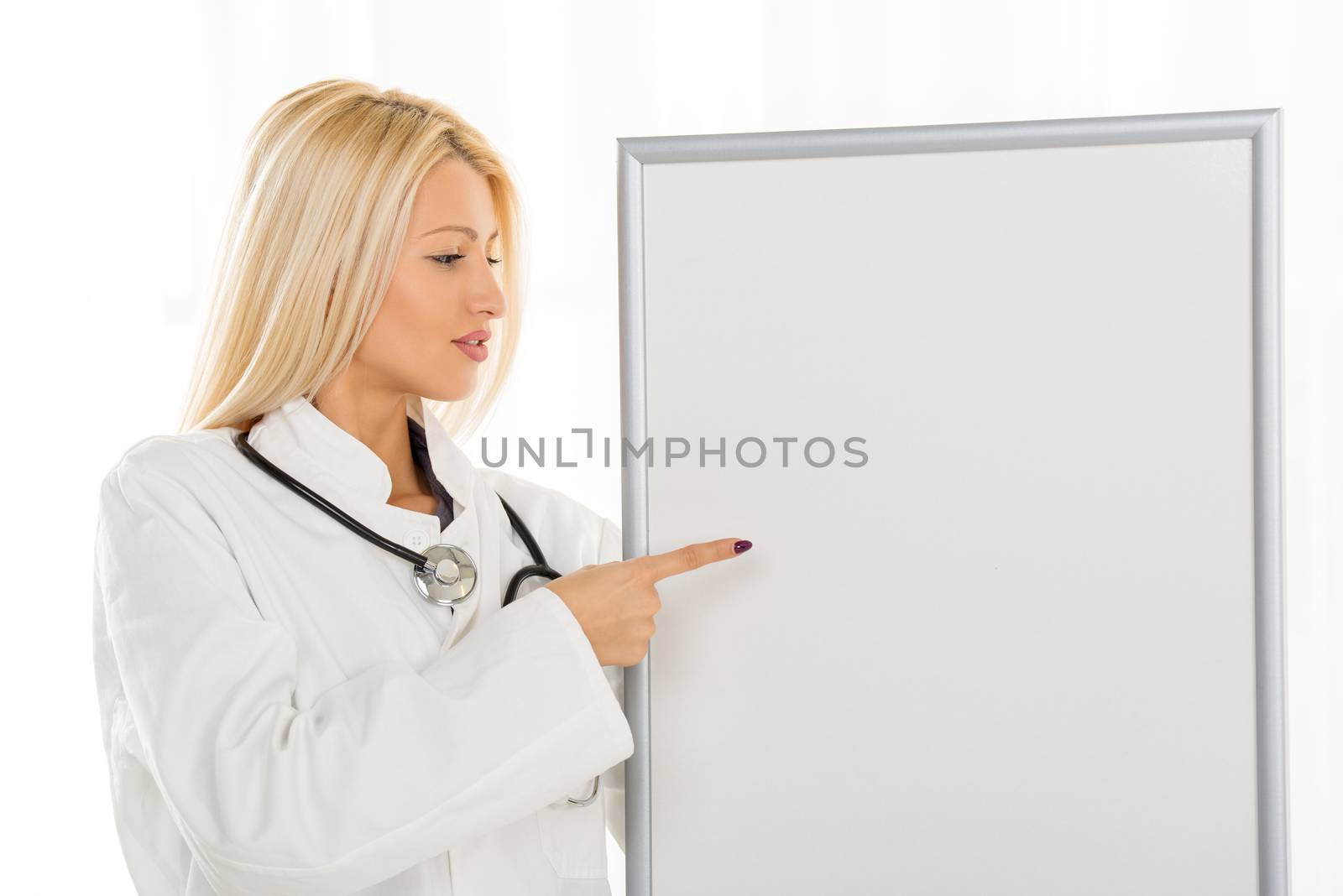 A pretty blonde girl in a white coat with a stethoscope over neck, standing in front of a table with a laptop, holding a blank notice board at which points a finger.