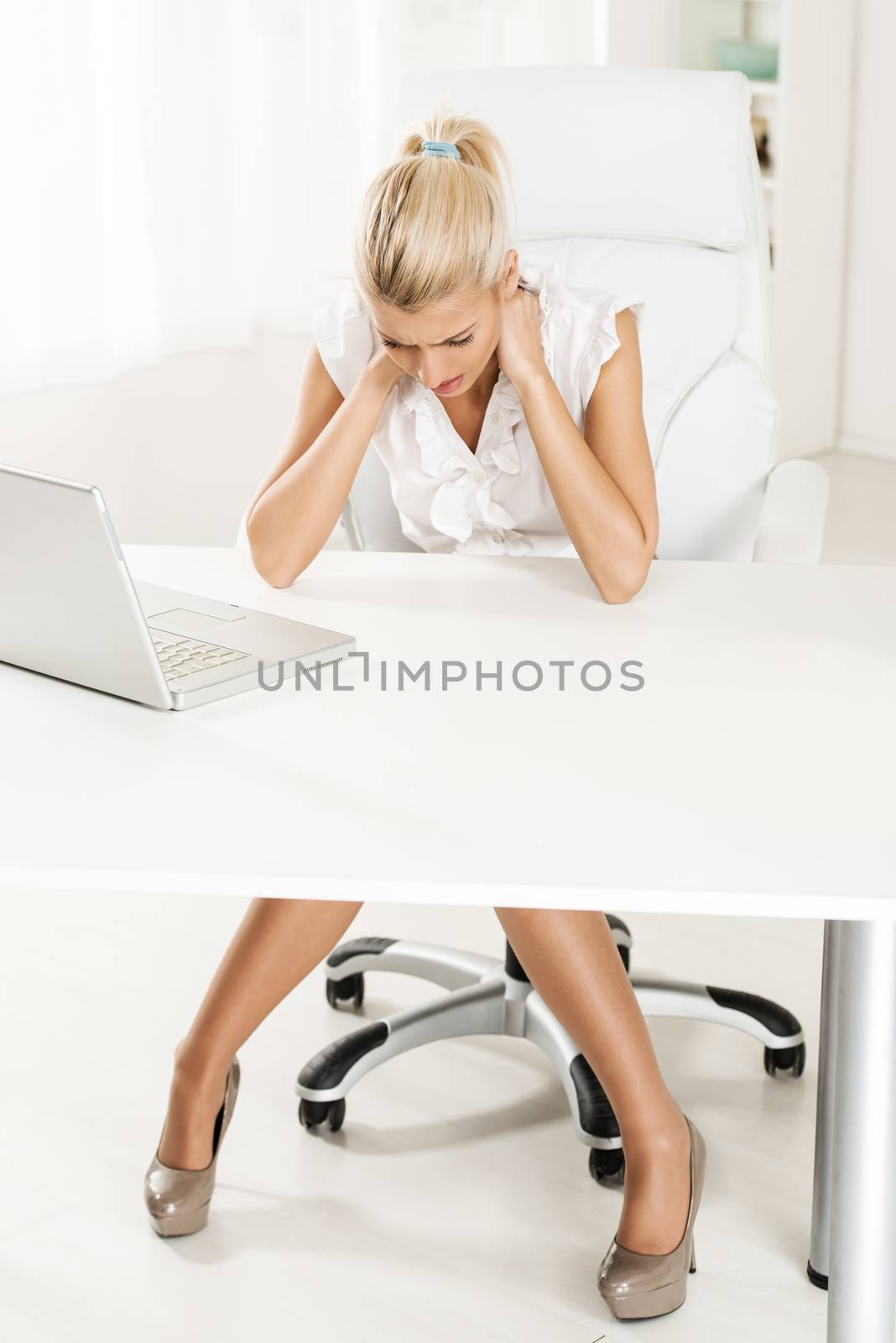 Young beautiful exhausted businesswoman sitting in the office, holding hands on the neck. In front of her on her office desk are laptop.
