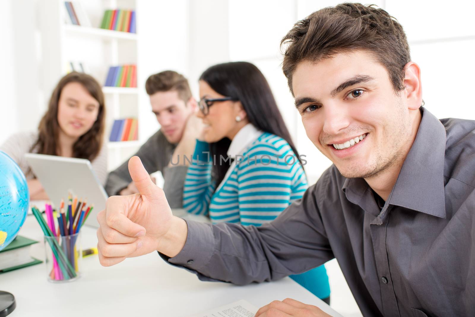 A beautiful smiling young man sitting and learning in the foreground. He showing thumb up and Looking at camera. A happy group of his friends is behind him and looking at laptop. 