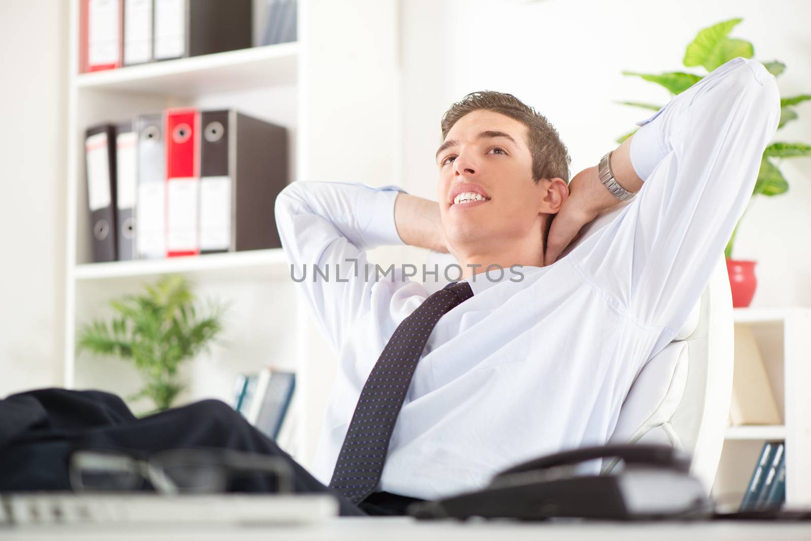 Young successful businessman relaxing in the office with hands behind  head and daydreaming. In the background you can see the shelves with binders.