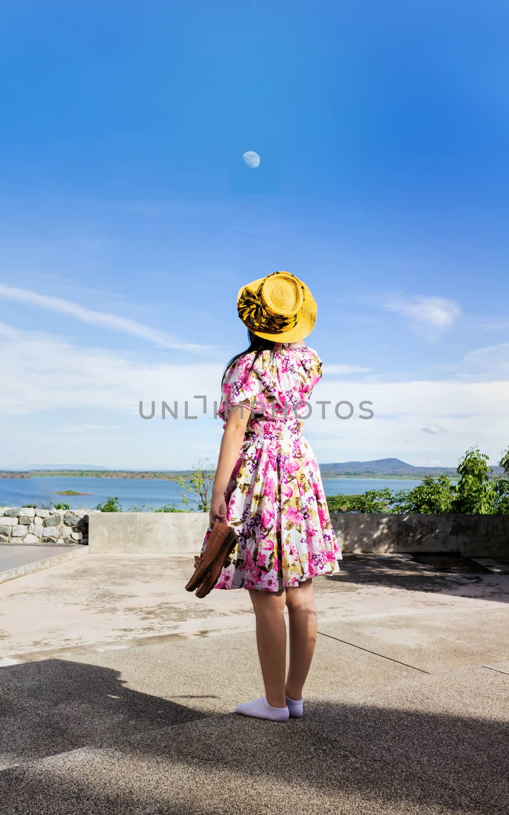 Women back or rare view standing and hold shoes look up to blue sky with little white moon, women or girl in summer flowers pattern dress with hat relax at reservoir or lake view background