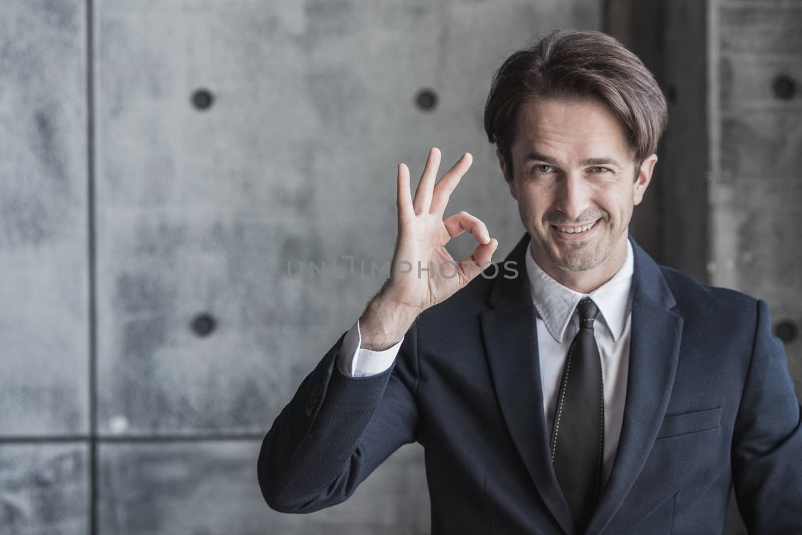 Portrait of businessman in suit showing ok sign over concrete wall background