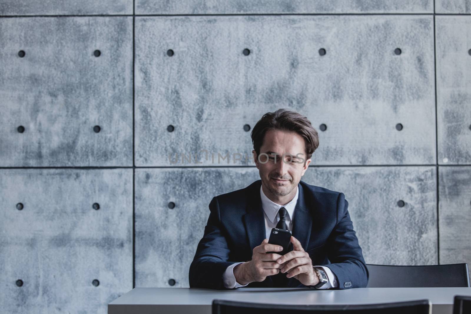 Portrait of a businessman with smartphone sitting behind the table over concrete wall background