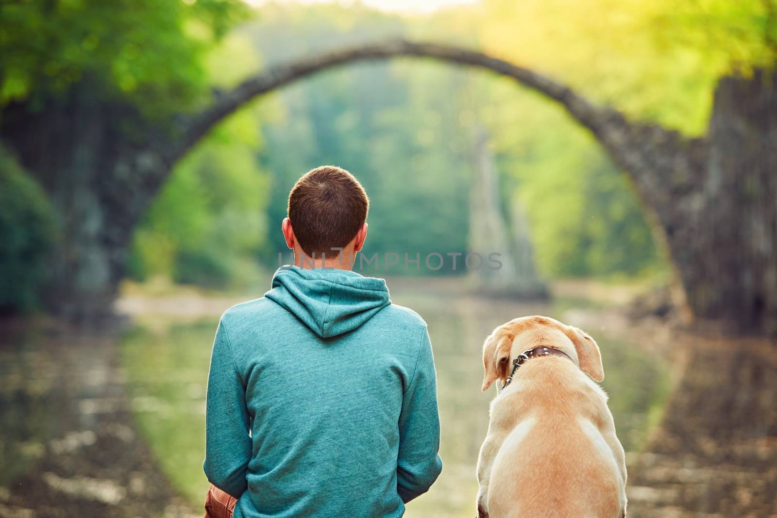 Pensive young man sitting on the pond bank with a his dog - Amazing place in Germany. Rakotzbrucke also known as Devils Bridge in Kromlau. 