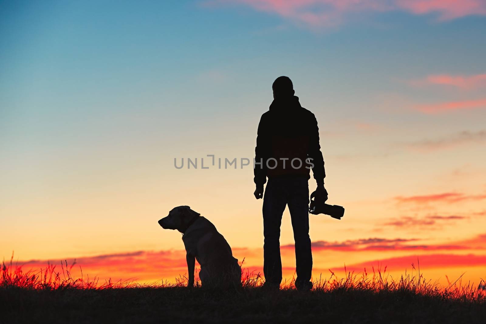 Silhouette of young photographer with his dog (labrador retriever) is enjoying sun. Photographer with mirror camera is ready to take sunrise pictures. 