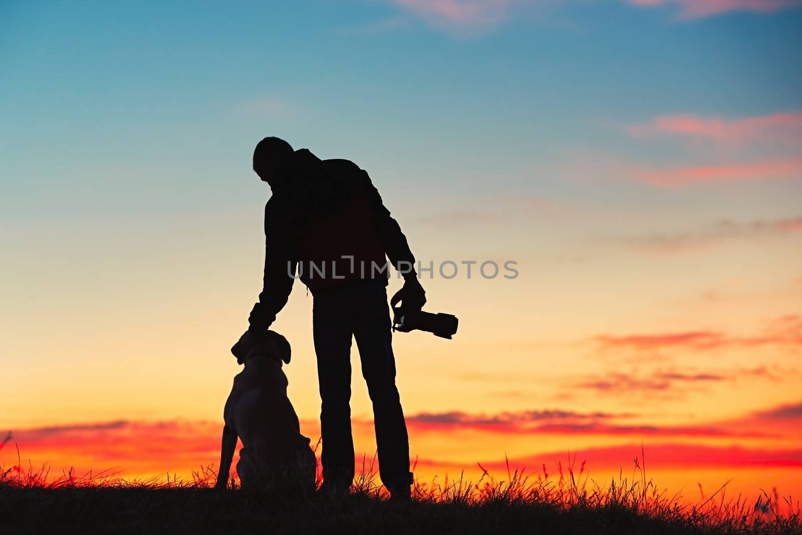 Silhouette of young photographer with his dog (labrador retriever) is enjoying sun. Photographer with mirror camera is ready to take sunrise pictures. 