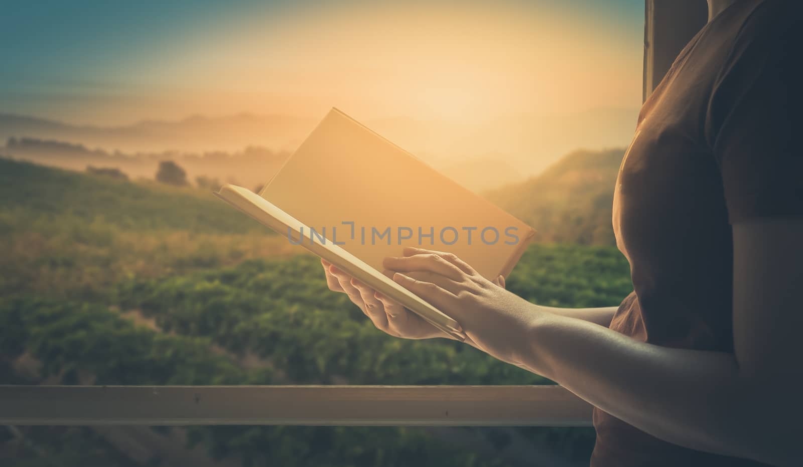 Young woman reading blank book beside window in morning time on weekend with mountain view in blurry background. Weekend lifestyle concept with vintage filter effect