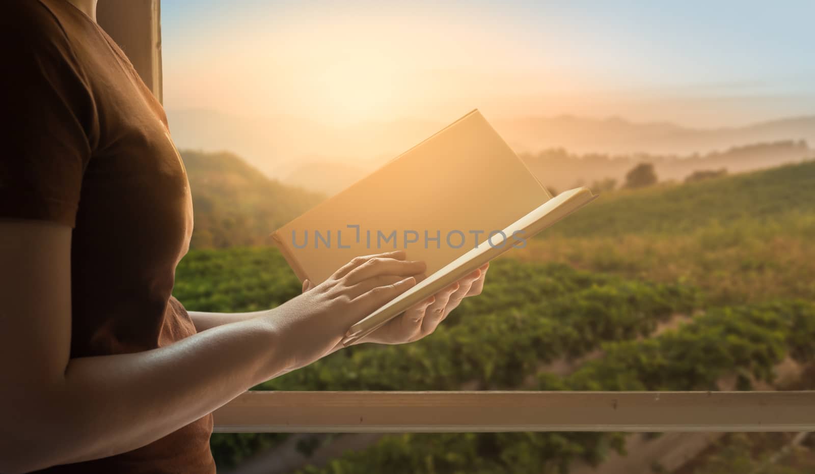 Young woman reading blank book beside window in morning time on weekend with mountain view in blurry background. Weekend lifestyle concept.