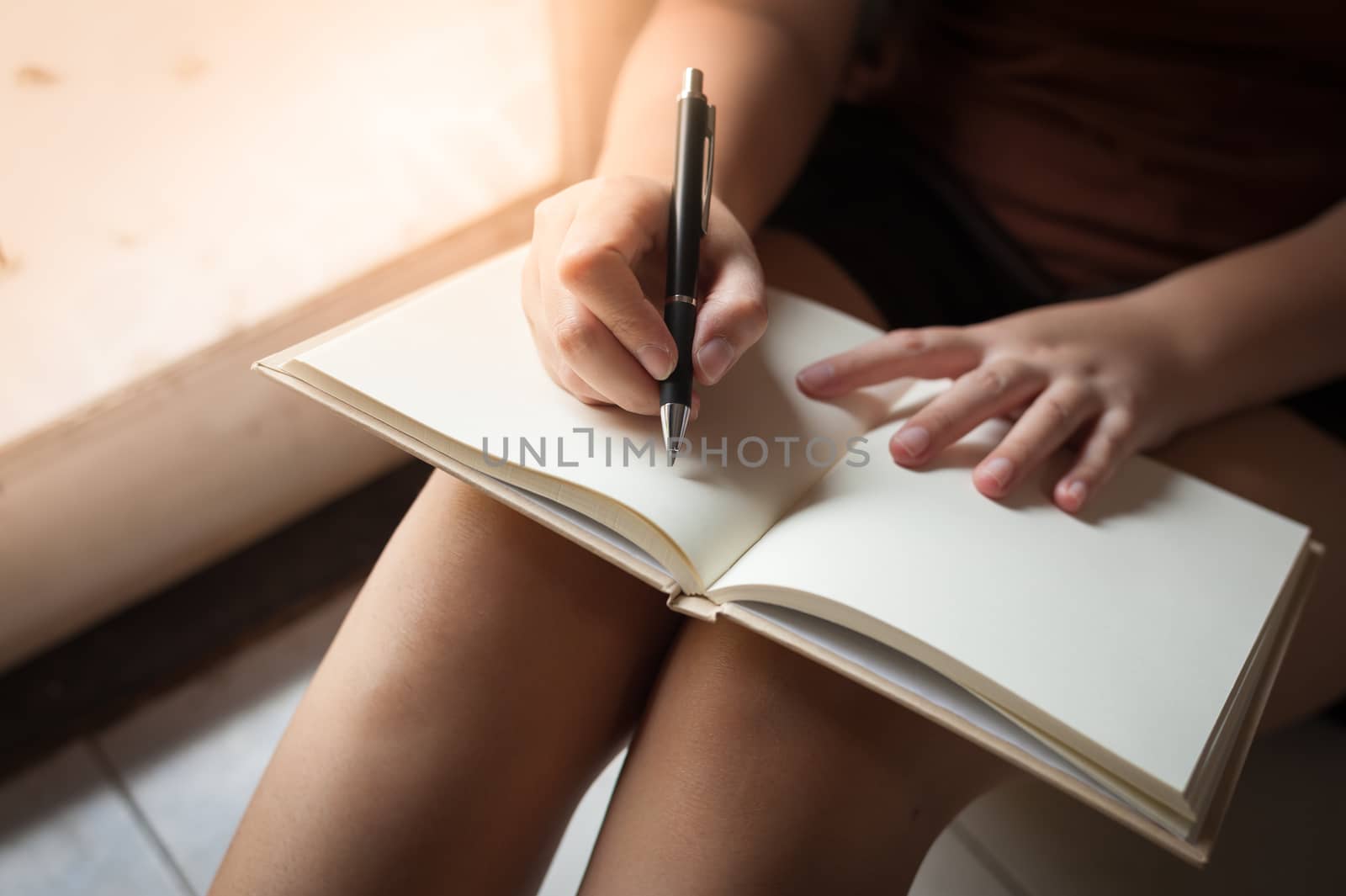 Young woman writing on blank book while sitting beside window in morning time on weekend. Morning lifestyle on weekend concept.