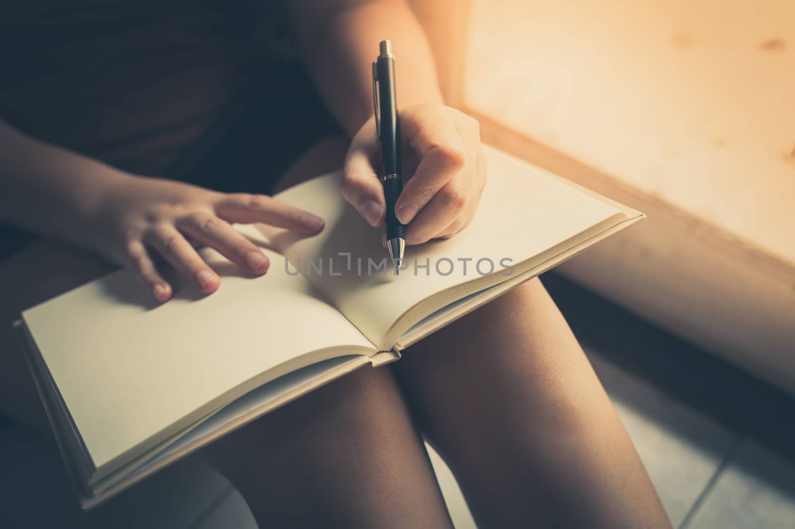 Young woman left hand writing on blank book while sitting beside window in morning time on weekend. Morning lifestyle on weekend concept with vintage filter effect