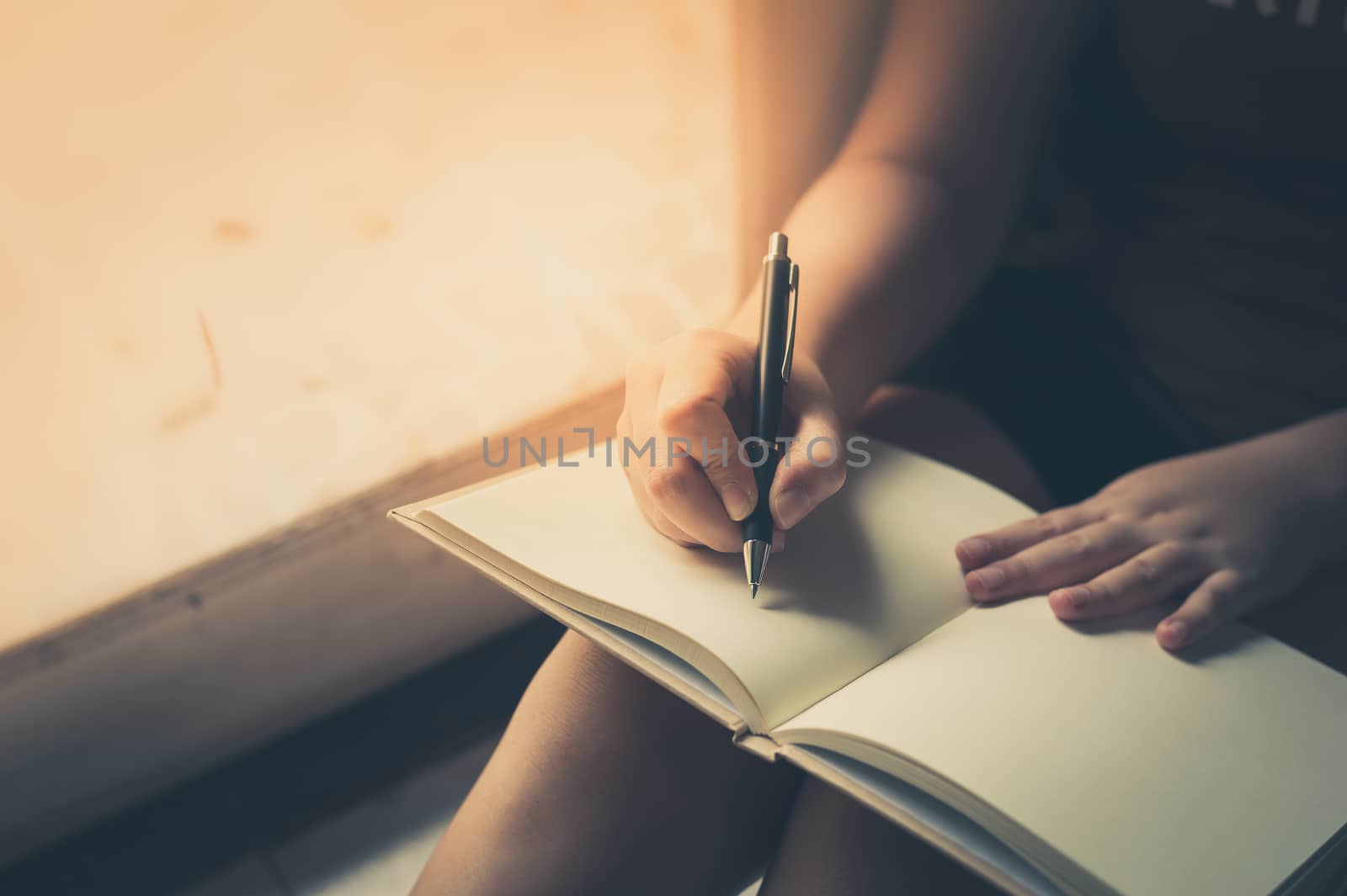 Young woman writing on blank book while sitting beside window in morning time on weekend. Morning lifestyle on weekend concept with vintage filter effect