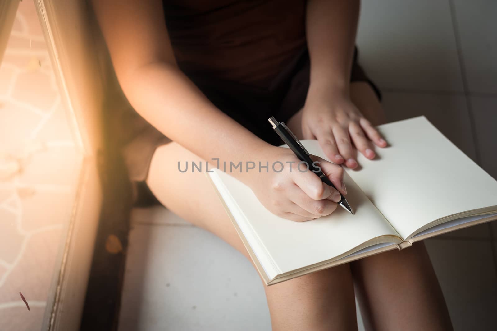 Young woman writing on blank book while sitting beside window in morning time on weekend. Morning lifestyle on weekend concept.
