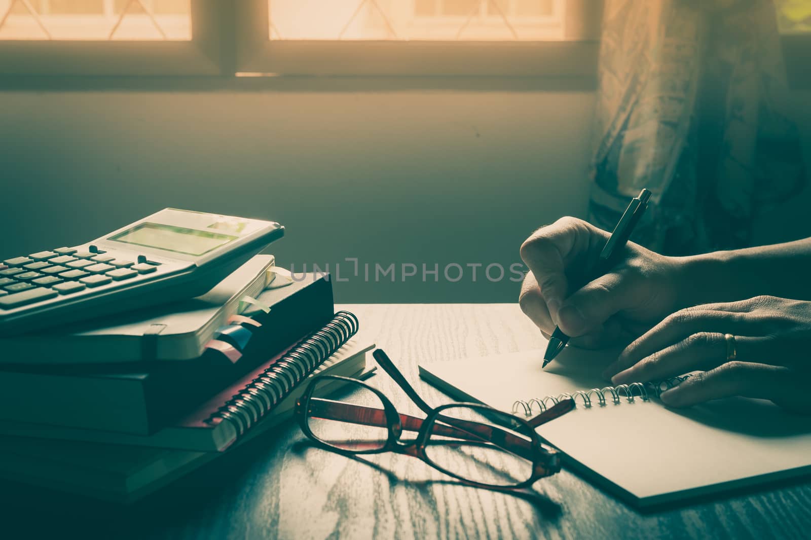 Male right hand writing on notebook beside glasses and books on table in morning time on work day. Freelance business working concpet with vintage filter effect