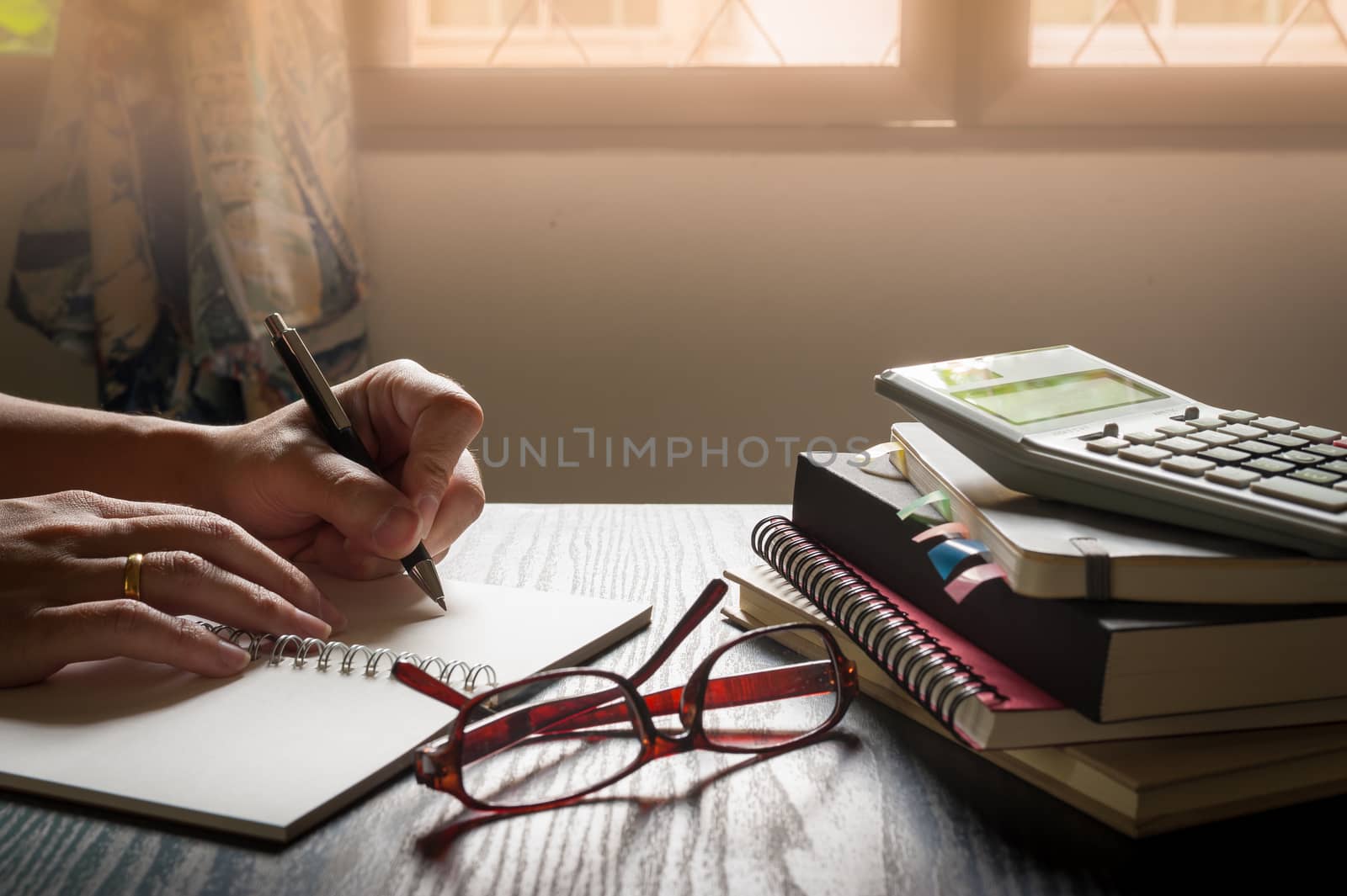 Male left hand writing on notebook beside glasses and books on table in morning time on work day. Freelance business working concpet