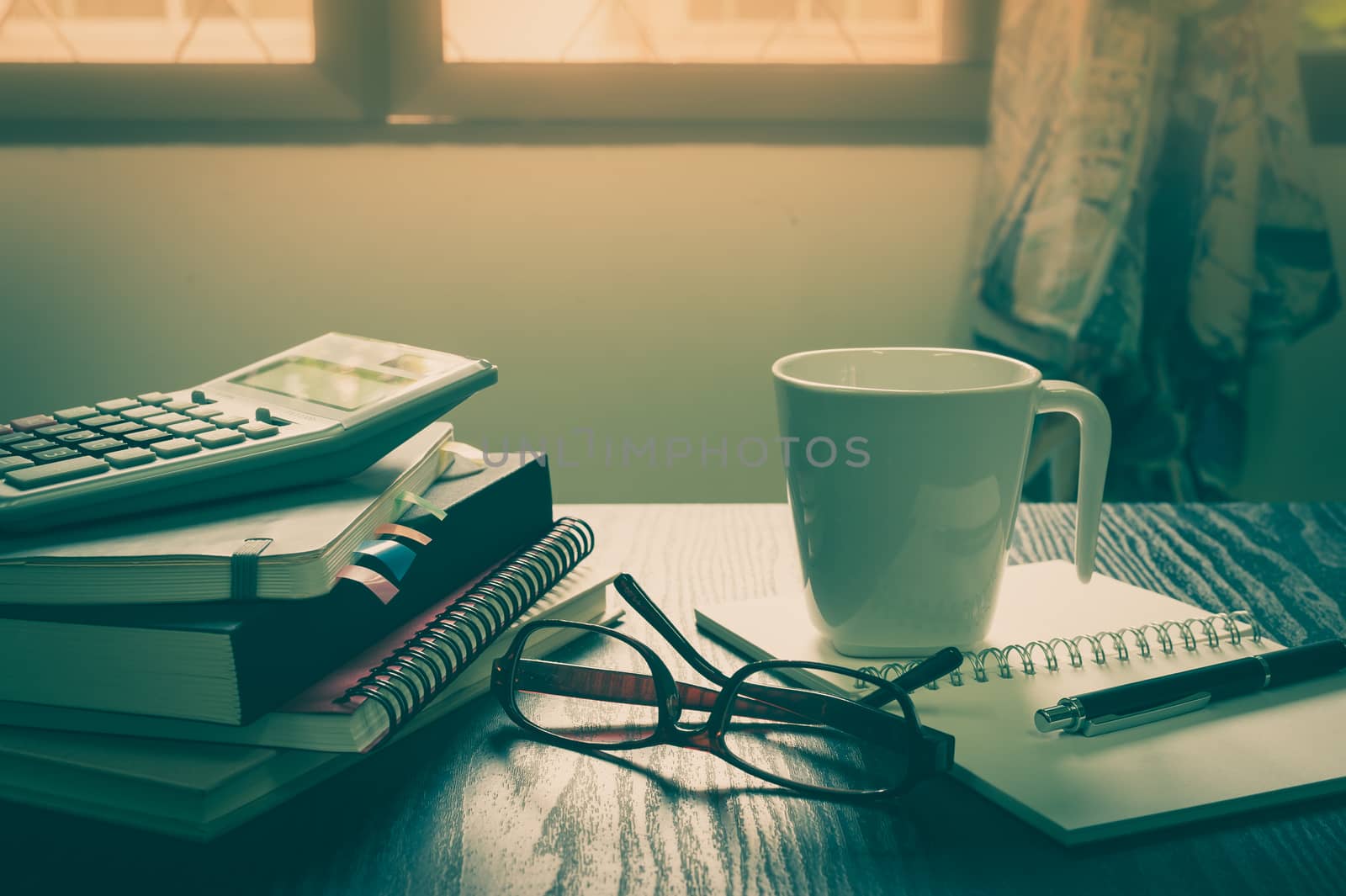 Coffee cup put down on table beside notebooks, glasses, and pen in morning time on work day. Freelance business working concept with vintage filter effect
