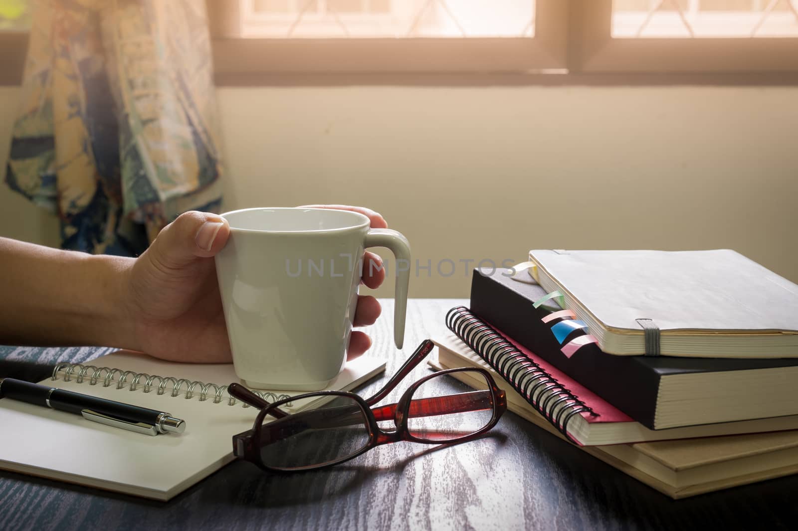 Young man left hand holding coffee cup on table beside notebooks, glasses, and pen in morning time on work day. Freelance business working concept