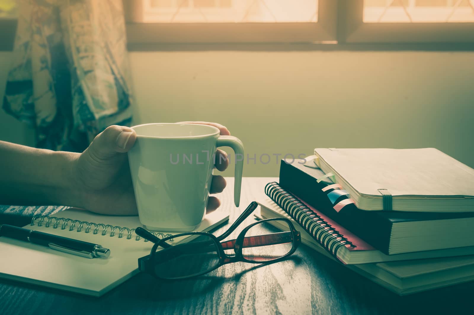 Young man left hand holding coffee cup on table beside notebooks, glasses, and pen in morning time on work day. Freelance business working concept with vintage filter effect