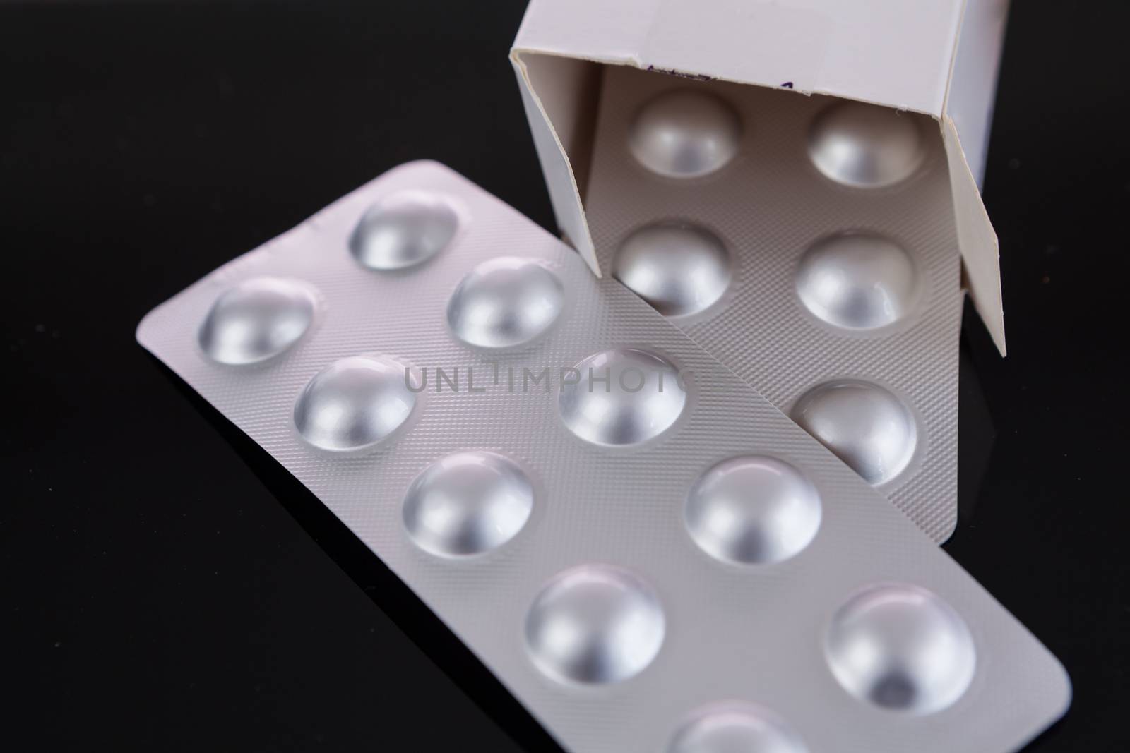 Silver blister pack of small pills together with the a box with additional packs showing through the open lid over a black background in a healthcare concept