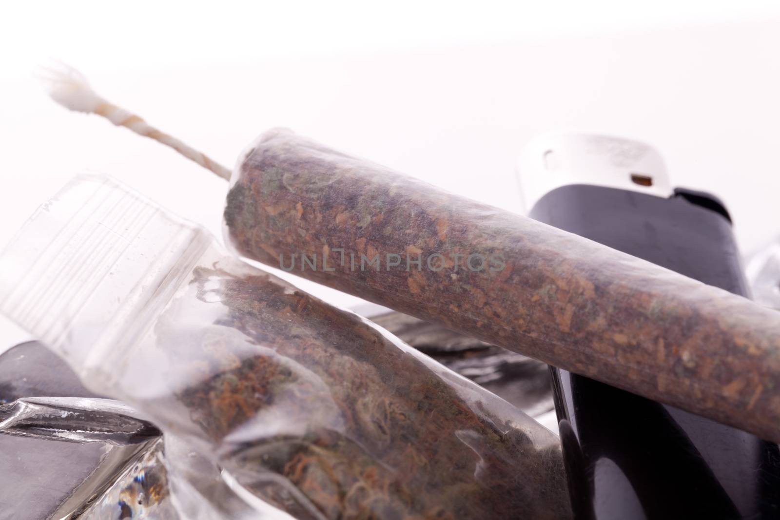 Close up of marijuana joint made with translucent rolling papers, plastic baggy of dried marijuana, black lighter and pipe on white background