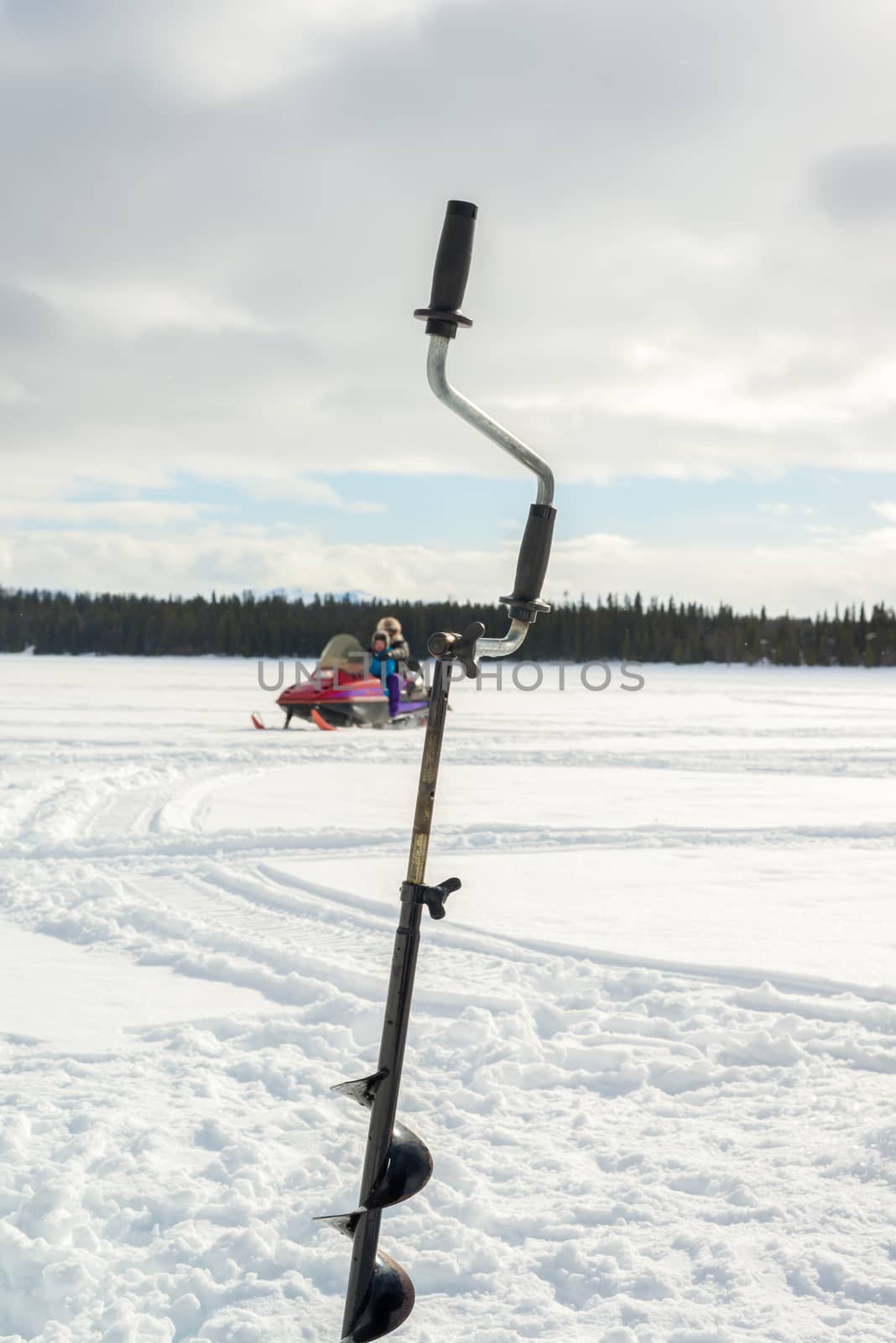 Scene with a ice fishing drill in the foreground while a parent and child is driving a snowmobile.