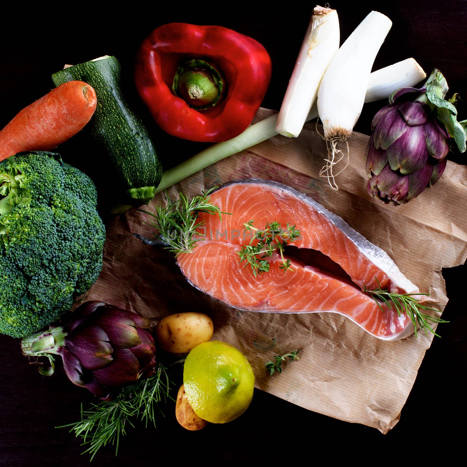 Raw Steak of Salmon and Heap of Raw Vegetables with Broccoli, Artichokes, Spring Onion, Potato, Red Bell Pepper and Lemon closeup on Parchment Paper. Top View on Dark Wooden background