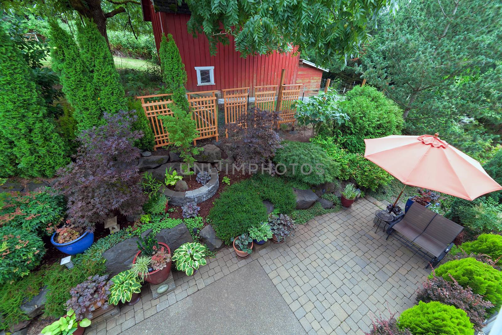 Backyard garden landscaping with paver bricks patio hardscape trees potted plants shrubs pond rocks furniture and red barn