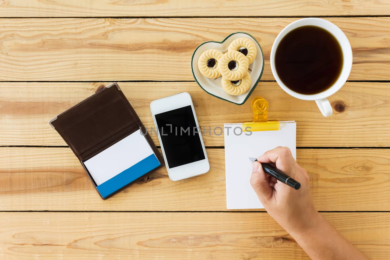 Top view hand with pen, writing down on blank paper note ,cellphone coffee and cookie on wood table