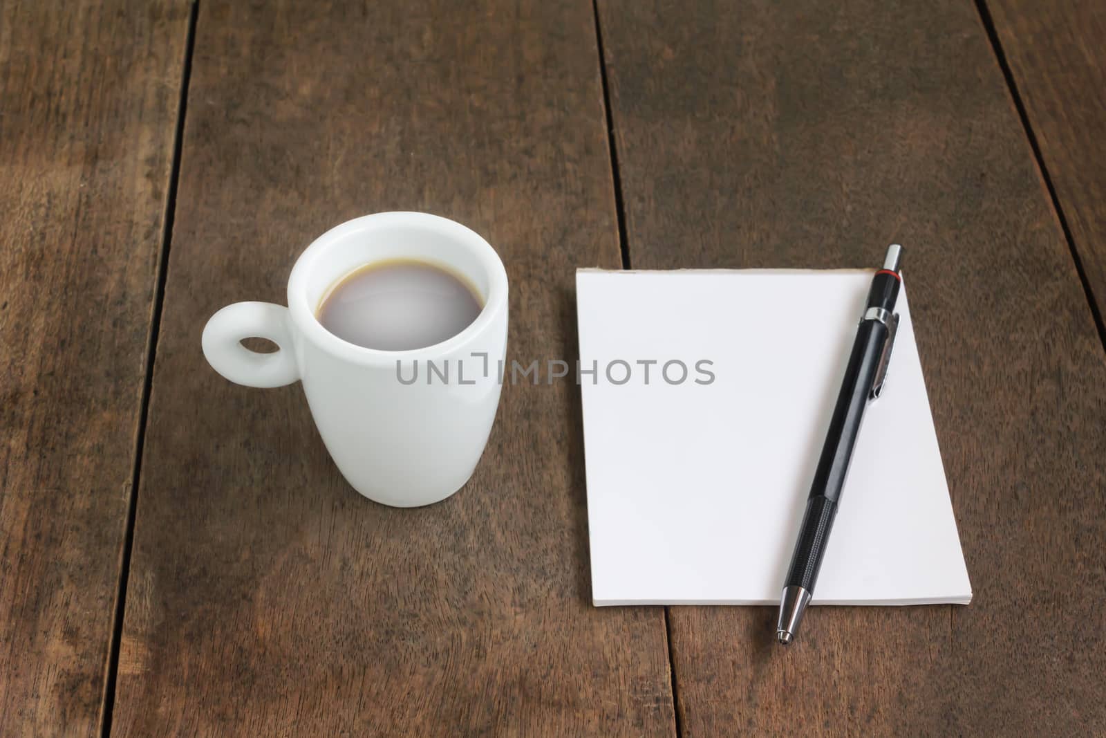 White cup of small hot black coffee with blank note book and pencil on wood table