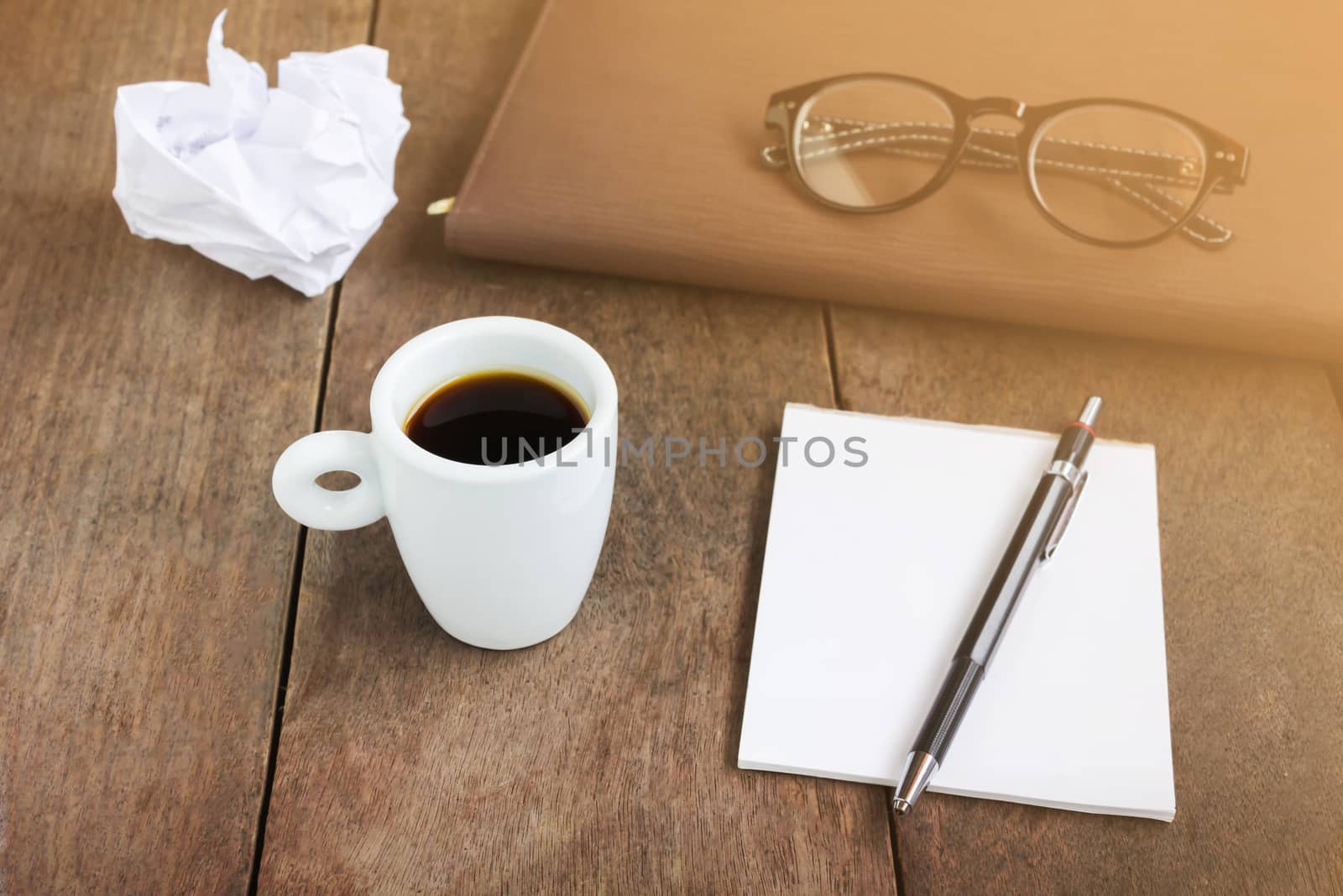 Vintage colour tone white cup of black coffee with blank note book and pencil with blurred eye glasses on wood table