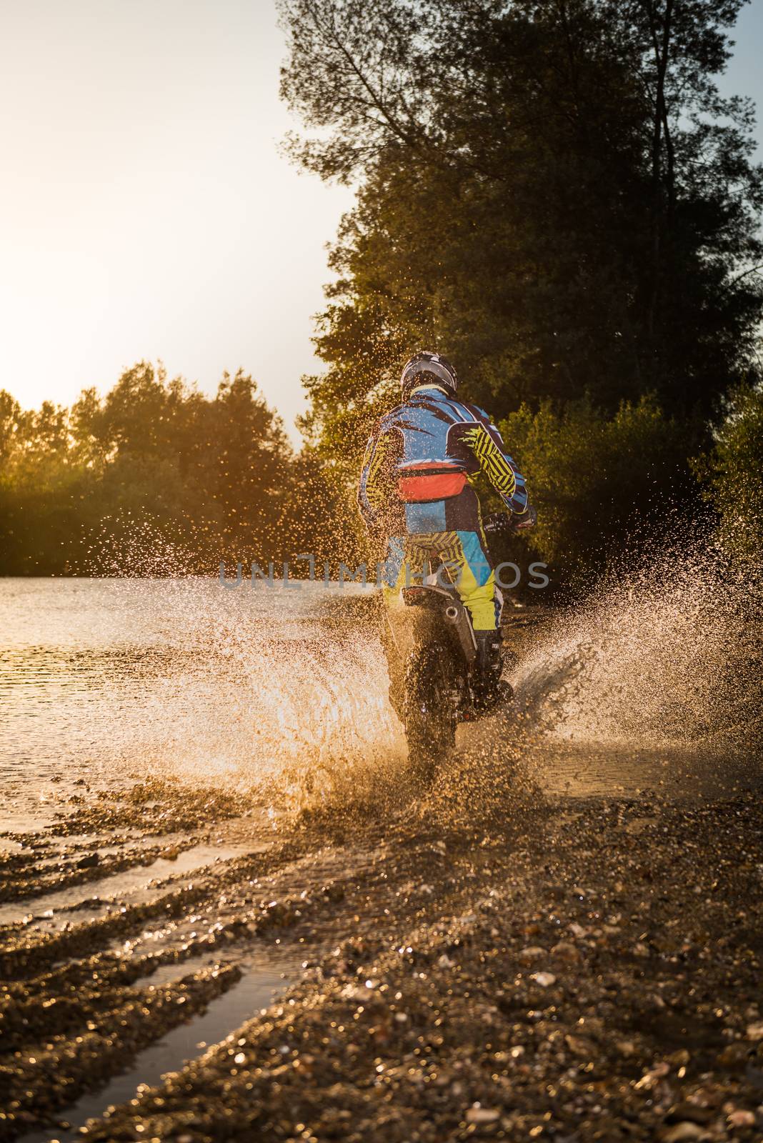 Enduro rider crossing water and muddy terrain against a beautiful sunset.