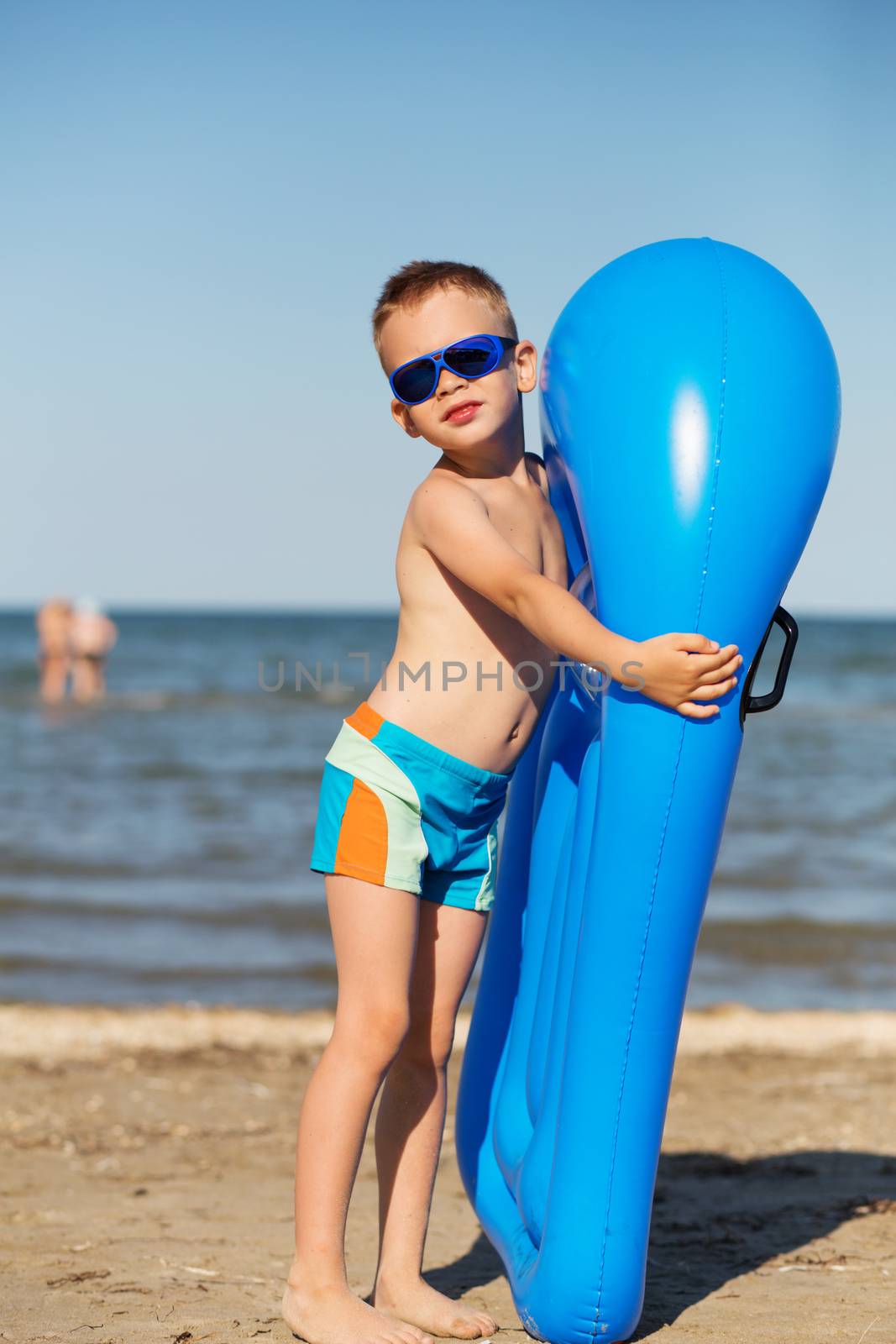 Little kid holding an inflatable mattress on the beach on hot summer day. Smiling boy playing on the beach with air mattress. Child is going to swim in the sea with swimming mattress. Summer vacation