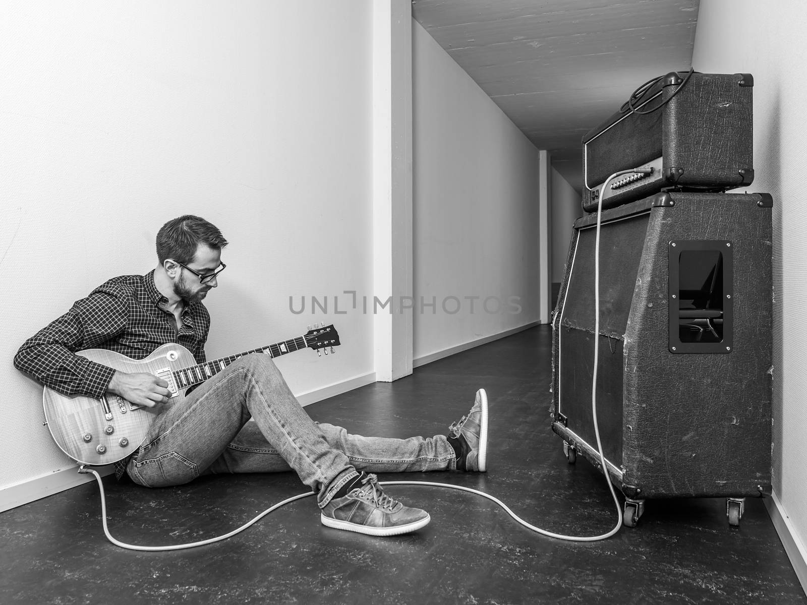 Photo of a man sitting playing his electric guitar in front of a large amplifier in a hallway. Black and white version.