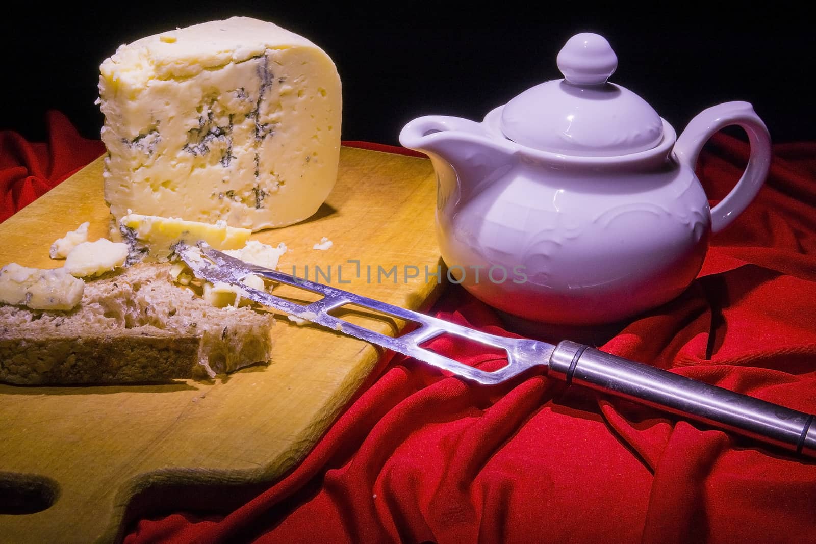 Triangular slice of Roquefort cheese is on the board next to the knife for cheese and white teapot