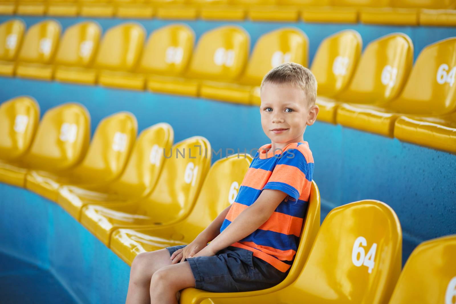 Little boy sitting in an empty stadium among yellow plastic numbered seats and applauds. Child take own seat in the stadium or dolphinarium and waiting patiently when the show or game starts.