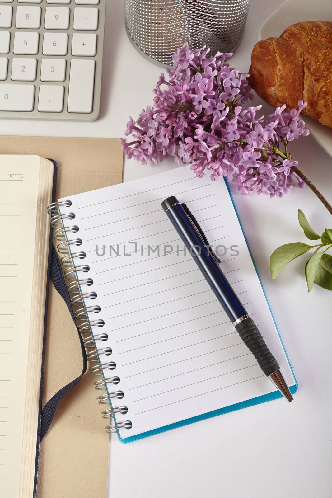 Top view office workplace - croissant, keyboard, calculator, pen, flower and notebook on white table