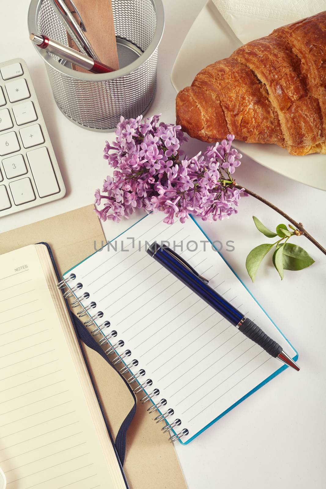 Top view office workplace - croissant, keyboard, calculator, pen, flower and notebook on white table
