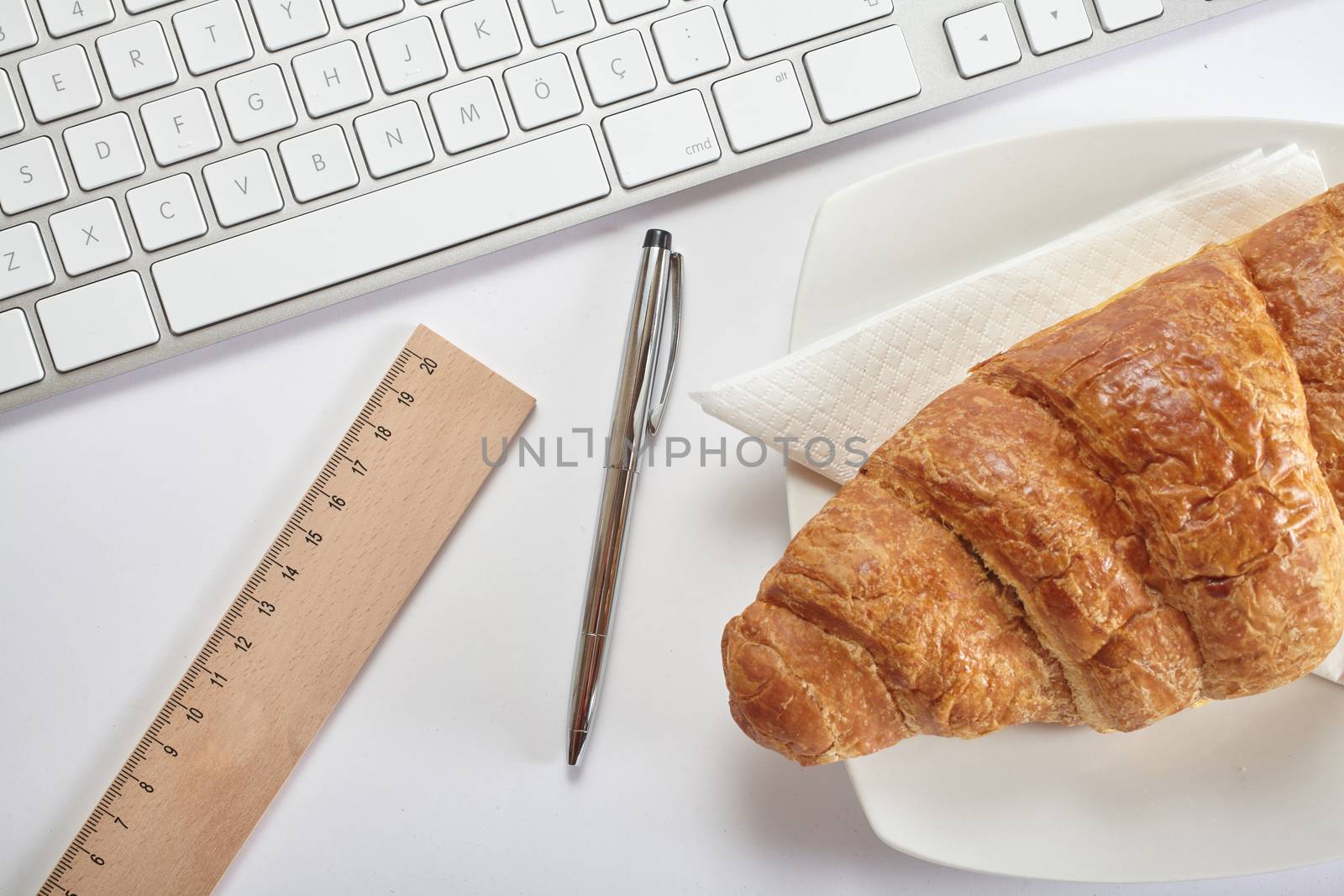 Top view office workplace - keyboard, pen, ruler and croissant on white table