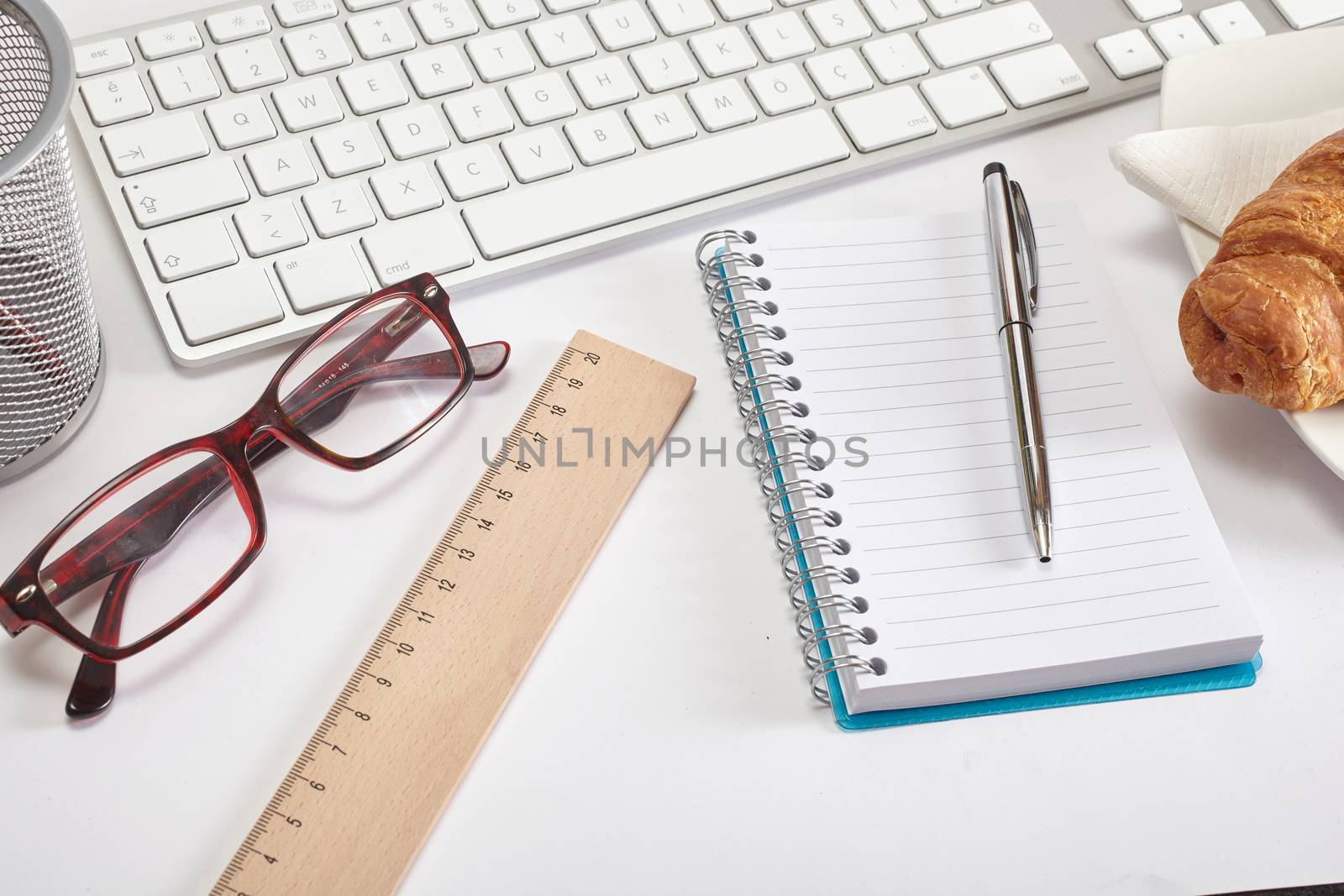 Top view office workplace - croissant, keyboard, pen, glasses, ruler and notebook on white table