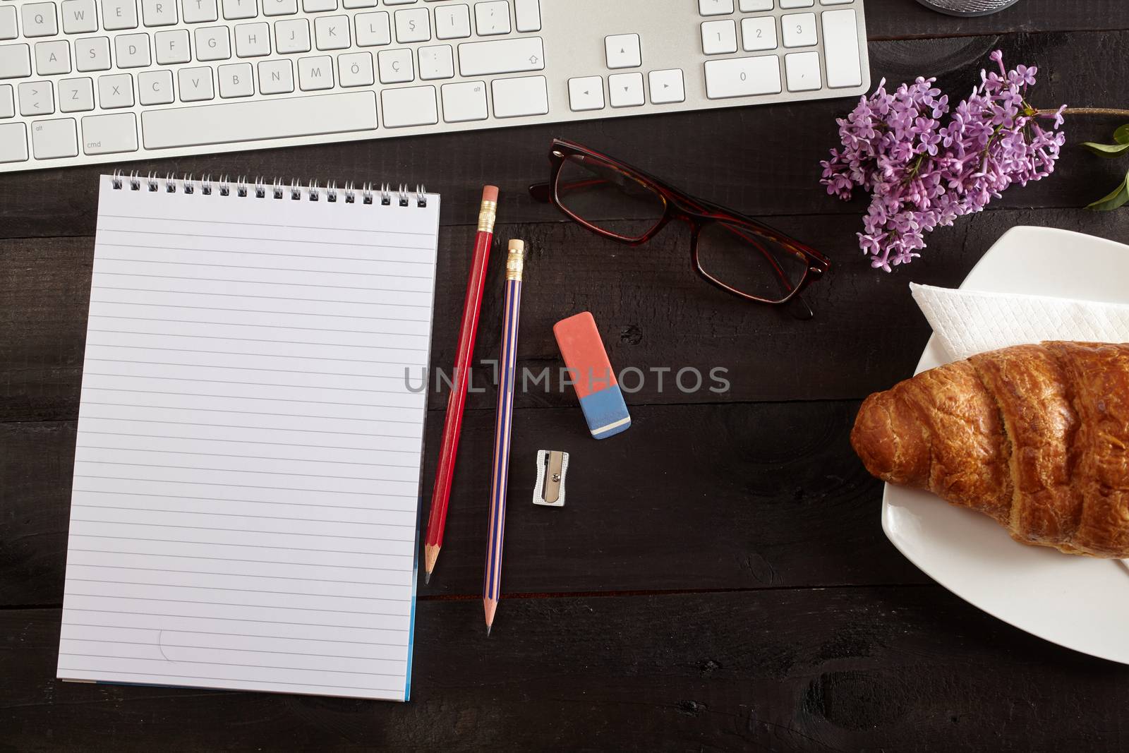Top view office workplace - keyboard, pencil, glasses, croissant and notebook on black table